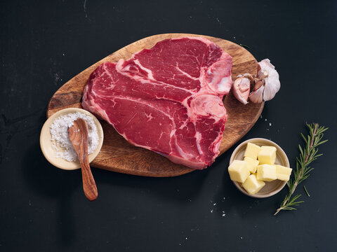 Raw t-bone steak on wooden cutting board with ingredients for cooking