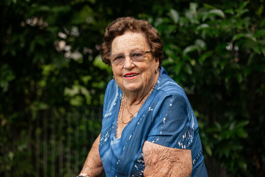 Aged woman standing outdoors in front of dense green foliage with metal fence behind
