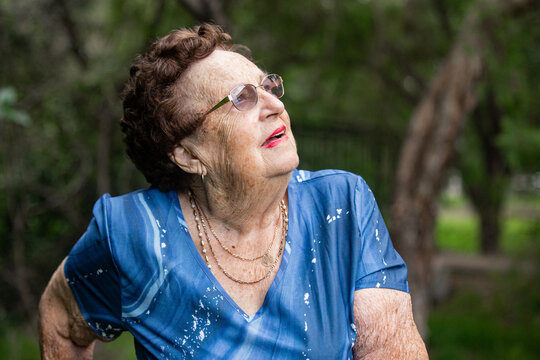 Aged woman wearing glasses, blue patterned shirt and gold necklaces looking up outdoors