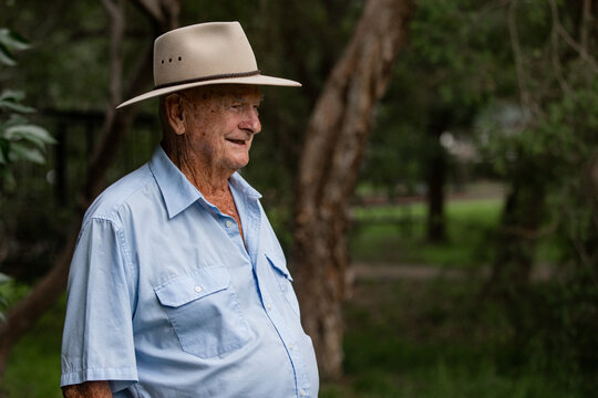 Australian old man in light blue button-up shirt and hat, standing outdoors among green trees