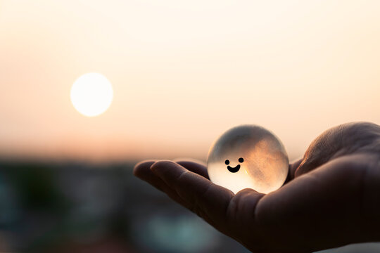 Hand holding glowing transparent sphere with fake smile against sunset. Concept of hidden depression, fragile mental health, emotional pain, inner wound, care, and healing hope.