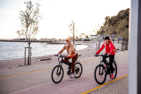 Two women friends riding electric bicycles by seaside path