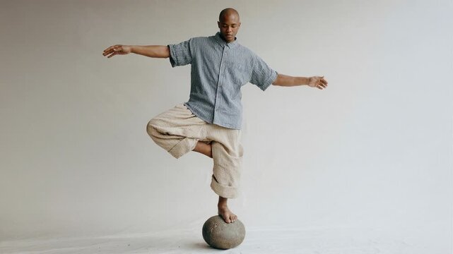 Man balancing on a stone sphere with arms outstretched in a studio setting. A focused individual demonstrates exceptional balance and control while standing on a spherical object