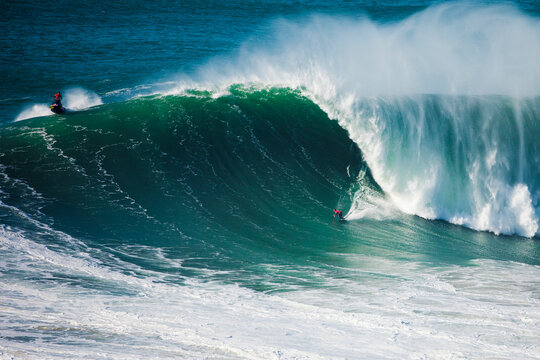 Surfer carving down a massive wave with spray and jet ski