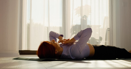 Fototapeta premium In a bright living room, a woman engages in stretching and pilates exercises on a mat. The sunny space promotes a healthy morning routine filled with movement.