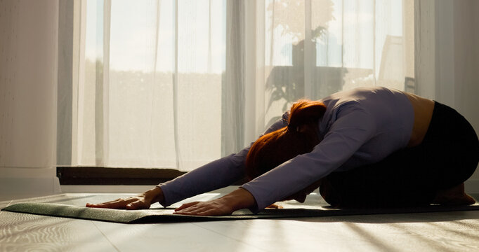 In a living room, a woman practices stretching and pilates on a mat, focusing on her fitness routine as the morning sun filters through the window.