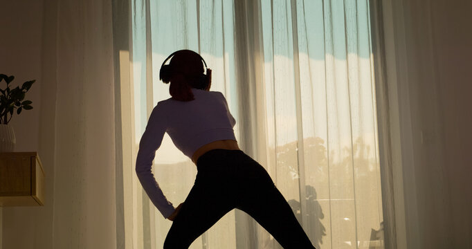 A woman enjoys a morning workout in her living room, dancing and exercising on the carpet while wearing headphones and feeling energized by the music.
