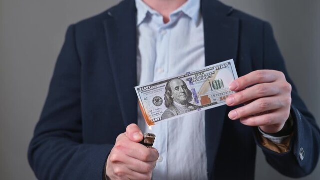 A man is holding a banknote as he ignites one edge with a lighter in a studio environment. The action takes place indoors, showcasing a dramatic moment with the burning money
