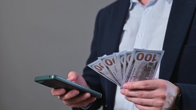 A man counts cash in his hands while holding a mobile phone in the other. He appears focused on managing his finances in a simple indoor space