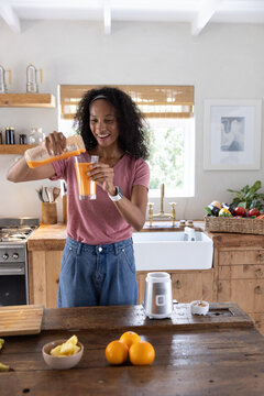 African American woman pouring orange juice from carafe into glass beside blender on kitchen island