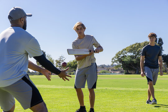 Three men playing cricket in sportswear on field, batter swinging flat wooden bat, ball airborne