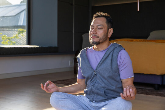 Middle-aged Asian man meditating cross-legged on floor in bedroom in hoodie, mustard throw nearby