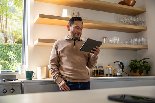 Mature Asian man wearing tan pullover, leaning in home kitchen and holding tablet