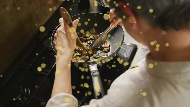 Kitchen cook stirring, flipping veg in pan with tongs, heat browning, gold dots partly hiding view