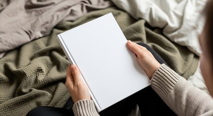 Woman's hands holding blank white book over cozy blankets in neutral tones