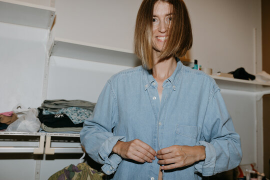 Smiling woman buttoning denim shirt while dressing at home