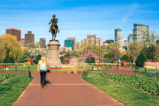 Motion blurred pedestrians in the Boston Public Garden near the George Washington statue, set against a backdrop of blossoming spring trees and the city's modern skyline