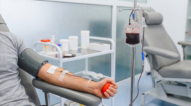 Man donating blood, with a full blood bag hanging from an IV stand, in a sterile medical clinic environment.