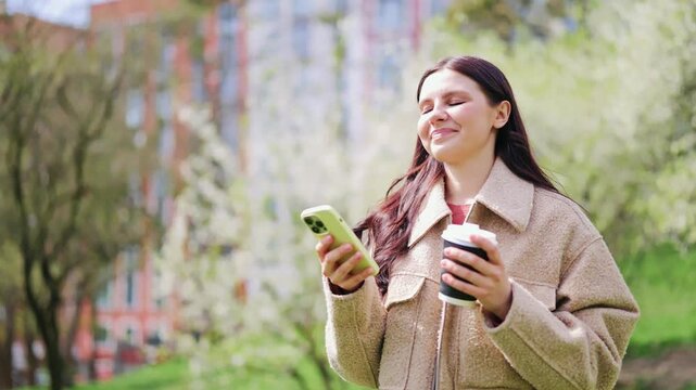 Smiling woman enjoys walk in park, holding phone and coffee. Feeling relaxed and happy. Nature s beauty enhances moment of contentment and connection.