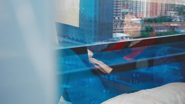 Woman at laptop wearing headset is typing on laptop filmed through balcony reflective glass at home workspace. Focused female Caucasian agent handles customer call and making notes. Freelance theme