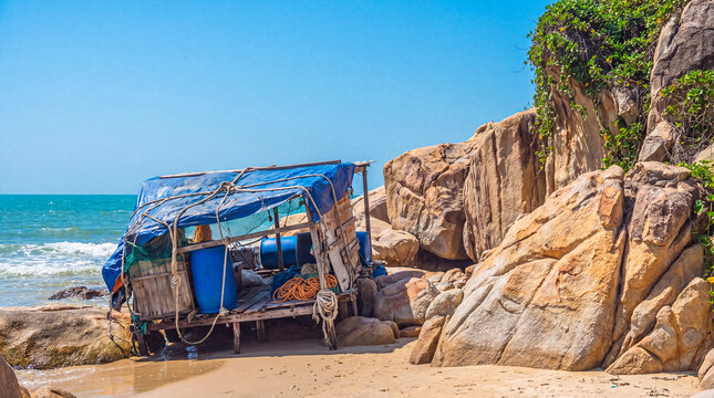 Makeshift fishing shack on a sandy beach by large rocks, with blue ocean and clear sky background.
