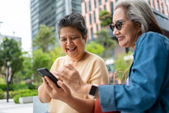 Two senior Asian women using phone shopping online outdoors in the city.