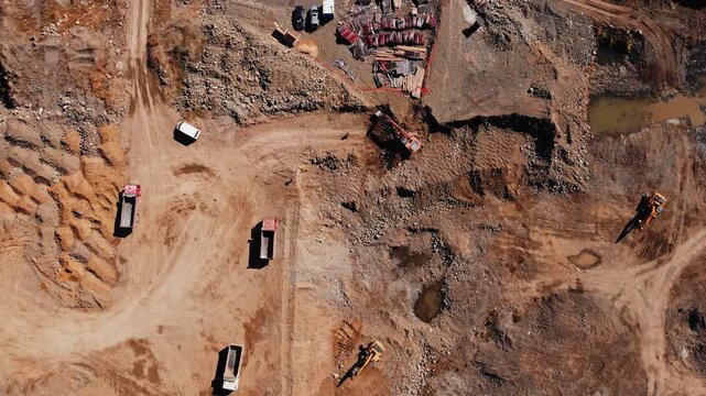 Multiple large construction vehicles are actively moving earth along dusty tires marks. Aerial view of excavation site with moving equipment. Multiple dump trucks are navigating dusty dirt tracks