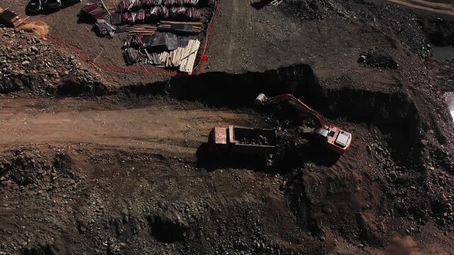 Aerial view of trucks and excavators navigating dirt lane with dust clouds during material delivery. Convoy of dump vehicles traverse compacted dirt lane. Top scene of construction site with vehicles.