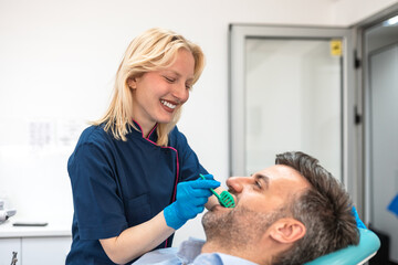 Fototapeta premium Smiling female dentist taking dental impression of male patient in clinic. Healthcare and medicine concept. Young woman doctor using tray for tooth mold, orthodontics or dental implant procedure.