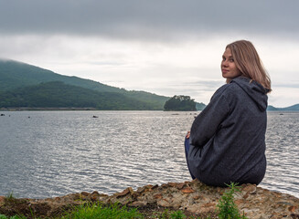 Fototapeta premium Young woman in grey hoodie relaxing by tranquil water with green mountains and small island under overcast sky