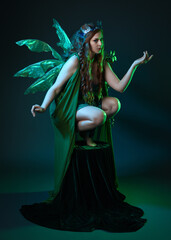 Fototapeta premium portrait of female model wearing fantasy fairy costume, green dress, cloak, butterfly wings and crown. full length crouching pose, isolated on dark studio background.
