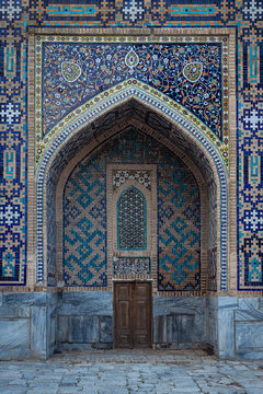 Ornate tiled iwan facade ща Tilla-Kari Madrasah with pointed arch and wooden door at Registan Square in Samarkand. Intricate Islamic mosaic patterns in turquoise and blue ideal for travel, heritage.