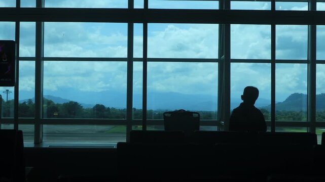 Silhouettes of children playing near chairs in a large airport waiting area before boarding, with a cloudy blue sky and views visible through panoramic glass windows. Makassar, Indonesia.