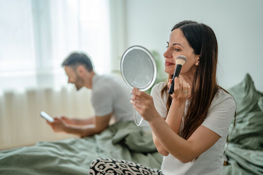 Woman doing makeup routine in bed while man uses phone