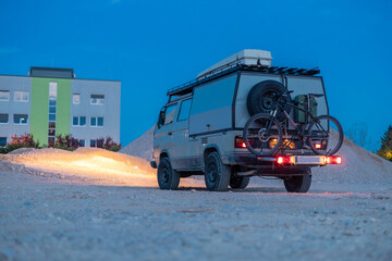 A rugged off-road camper van parked on gravel at dusk, equipped with a roof rack, mountain bike, and spare tire. A modern green-accented building stands in the background under a blue twilight sky. © Anze