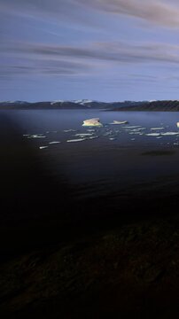 Icebergs float in a wide body of water during dusk. The sky shows soft colors as the sun sets behind the mountains in the background. This scene captures the beauty of nature at twilight.