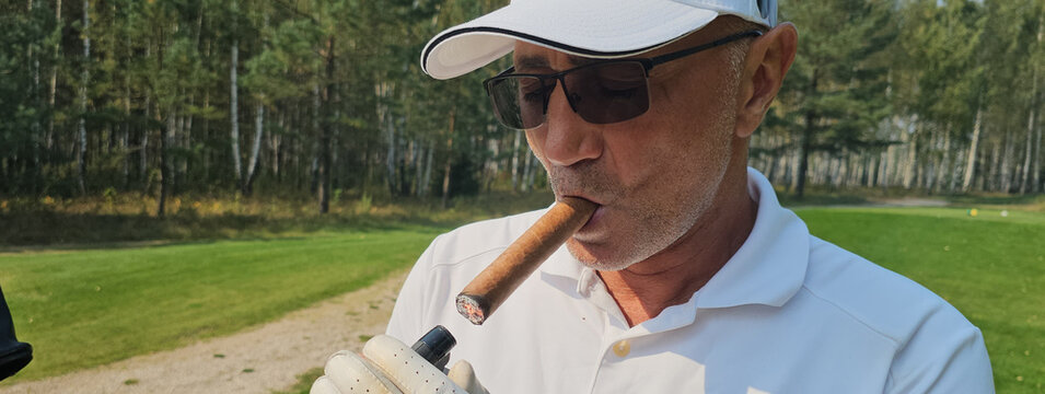 A golfer enjoys a cigar while relaxing on the course amidst green trees during a sunny day in early autumn