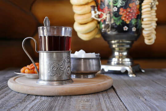 On a wooden table on the background of log wall there is a glass of black tea in a metal cup holder next to a samovar with bagels. 
