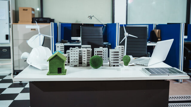 Modern office desk with solar panel, wind turbine models, hard hat, eco house and city miniatures, laptop, emphasizing renewable energy, sustainability, and green architecture planning.