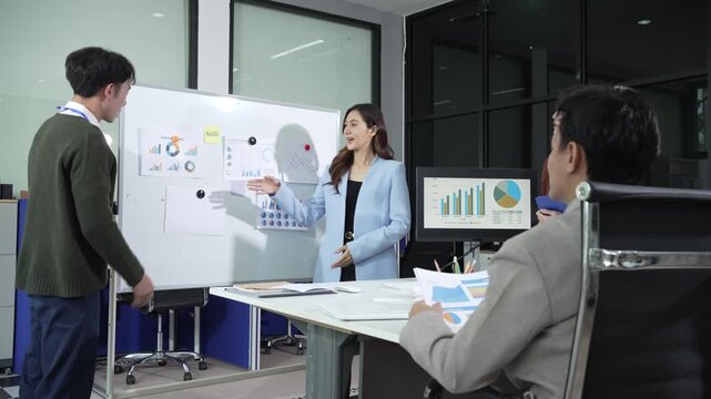 Business team meeting in modern office with woman presenting charts on whiteboard, colleagues reviewing printed reports and monitor with graphs, corporate strategy and data analysis concept