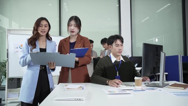 Office teamwork scene: two female managers reviewing documents beside a male employee working at desktop computer in modern open-plan workplace.