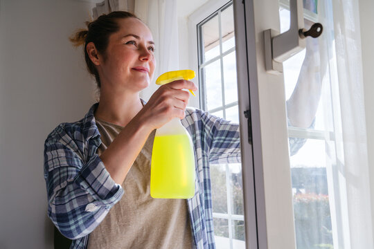 Smiling woman cleaning window with spray detergent at home