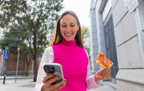 Smiling woman eating pizza and using smartphone on a city street