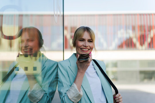 Happy architect talking through mobile phone speaker leaning on glass wall