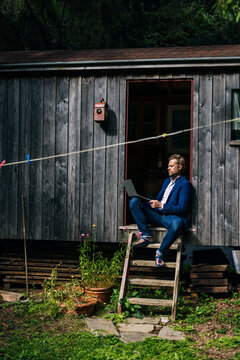 Mature businessman using laptop and sitting on doorway of log cabin