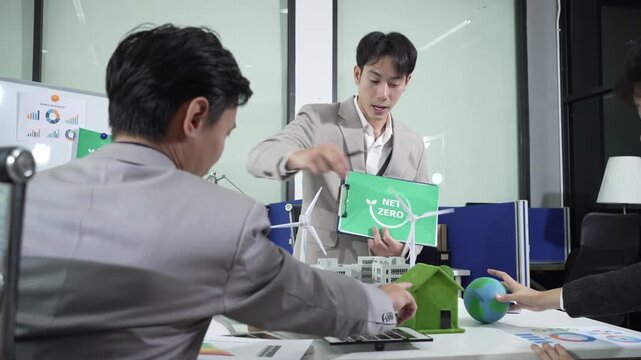 Business meeting about sustainability; team reviews net zero sign with wind turbine model, eco house, and globe in modern office, discussing green energy strategy and emissions reduction.