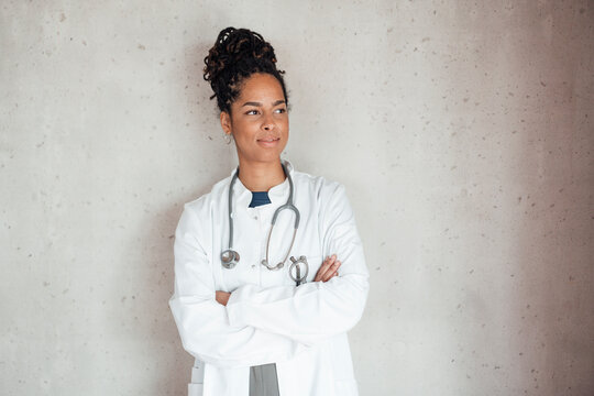 Smiling young female doctor with arms crossed leaning on gray wall at hospital