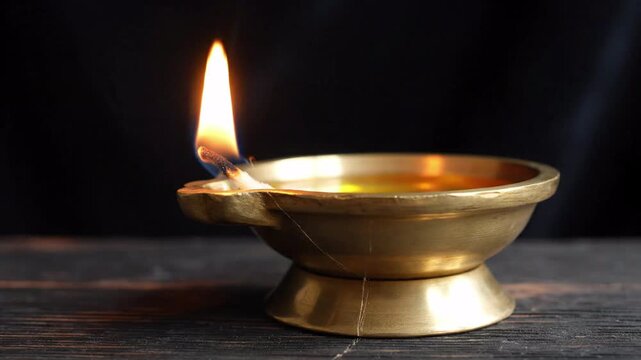 Close up of an oil lamp with a lit wick and flame, burning oil in a brass vessel, on a dark rustic wooden surface with a black background