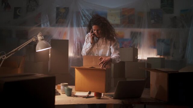 Concerned female small business owner talking on phone while packing cardboard boxes for shipping in office warehouse.