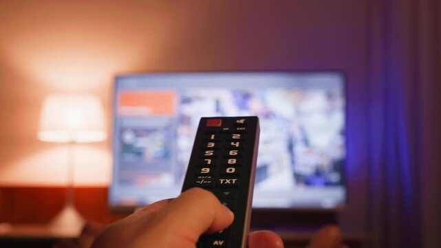 A close-up of a hand holding a TV remote control and changing channels. The TV screen is blurred in the background, creating a modern concept of home entertainment and relaxation. High quality 4k